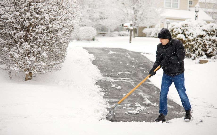 A person shovels snow from a residential driveway that is surrounded by shrubs and trees.