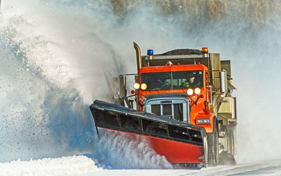 A snow plow causes a plume of salt-treated snow to spray up into the air, potentially causing salt damage to surrounding trees and plants.