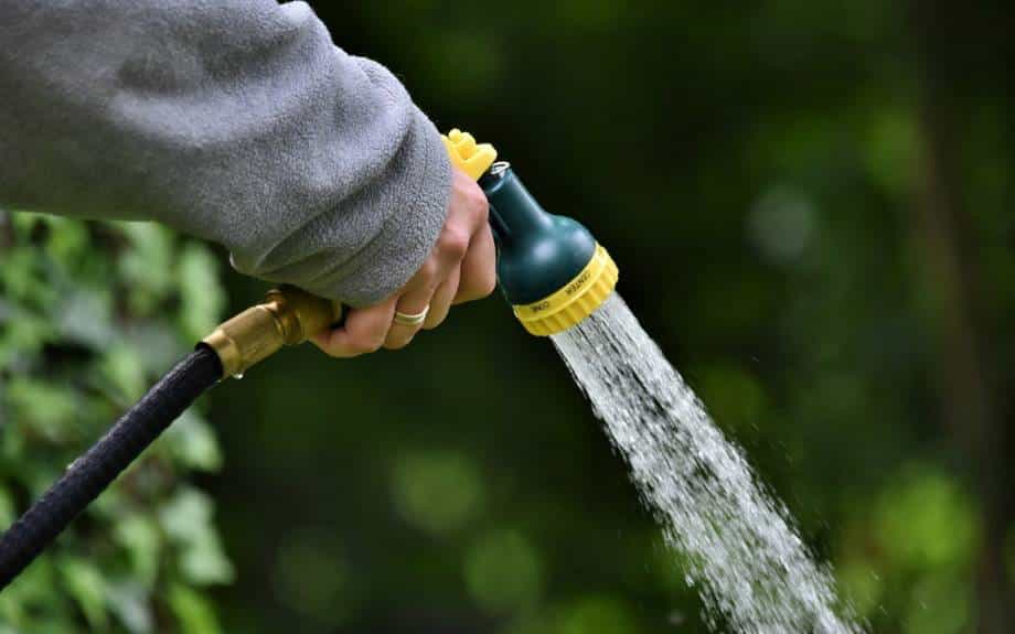 A hand with a gold ring on a finger pokes through the armhole of a gray sweatshirt and clutches a water hose attachment as it sprays water toward the ground with green foliage in the background.